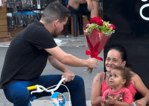 Homem entrega uma bicicleta infantil e flores a uma mãe sentada no calçadão de Osasco, enquanto seu filho pequeno sorri ao experimentar a bicicleta, em um gesto de empatia, cuidado e reconhecimento materno.